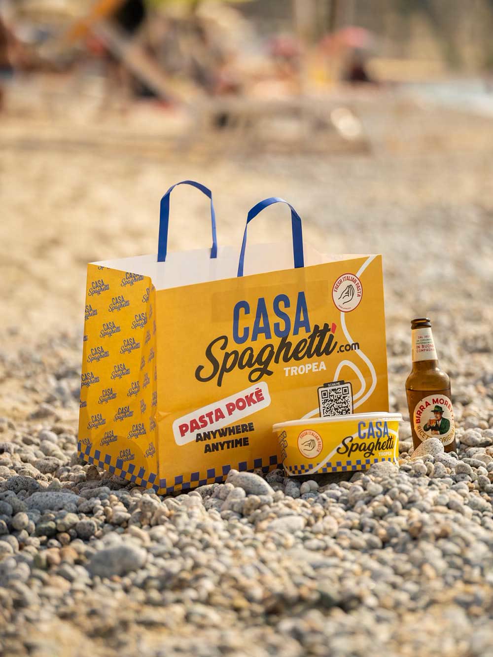 La borsa Casa Spaghetti sui ciottoli della spiaggia, con la ciotola e la birra